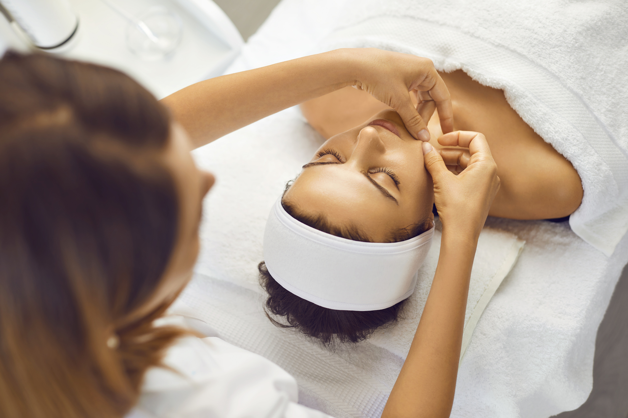 Cosmetologist making facial massage for serene young womans face with hands in beauty spa salon, top view. Facial treatment, massage, skincare, cosmetology concept
