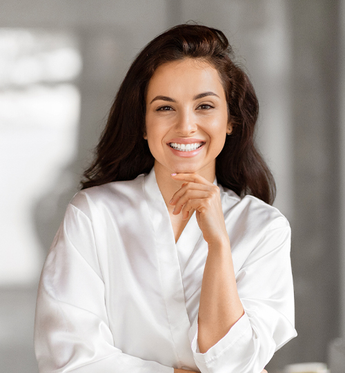 Smiling woman in white blouse with hand on chin, natural lighting portrait in a clean indoor setting