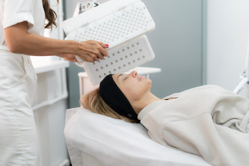 Client undergoing LED light therapy facial with overhead panel device in a clinical skincare treatment room