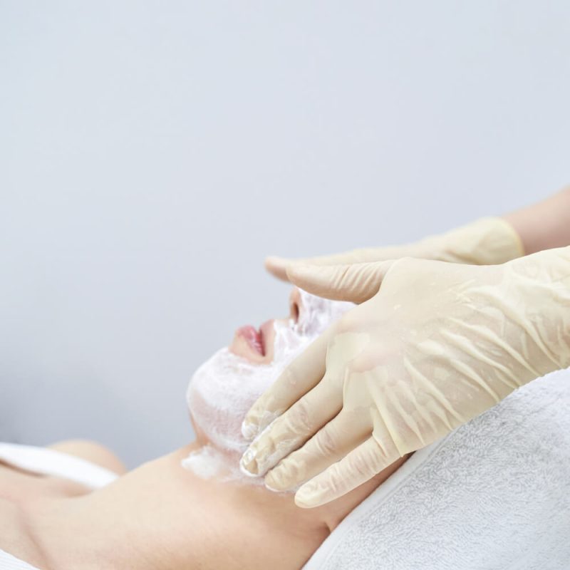 Aesthetic therapist applying facial cleanser or mask to client’s face using gloved hands in a clinical spa setting